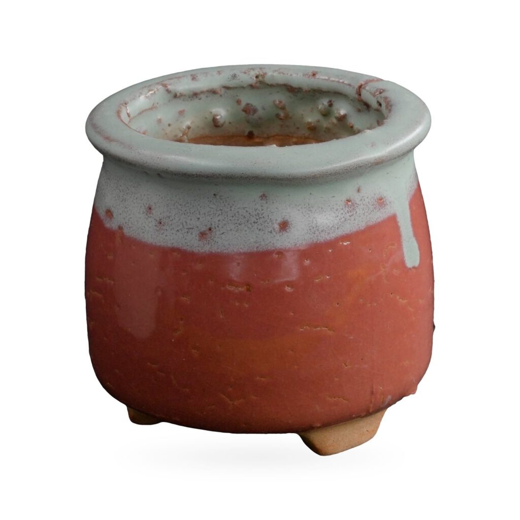 Small round footed pot in glossy coral red with pale celadon-green glaze dripping from the rolled rim